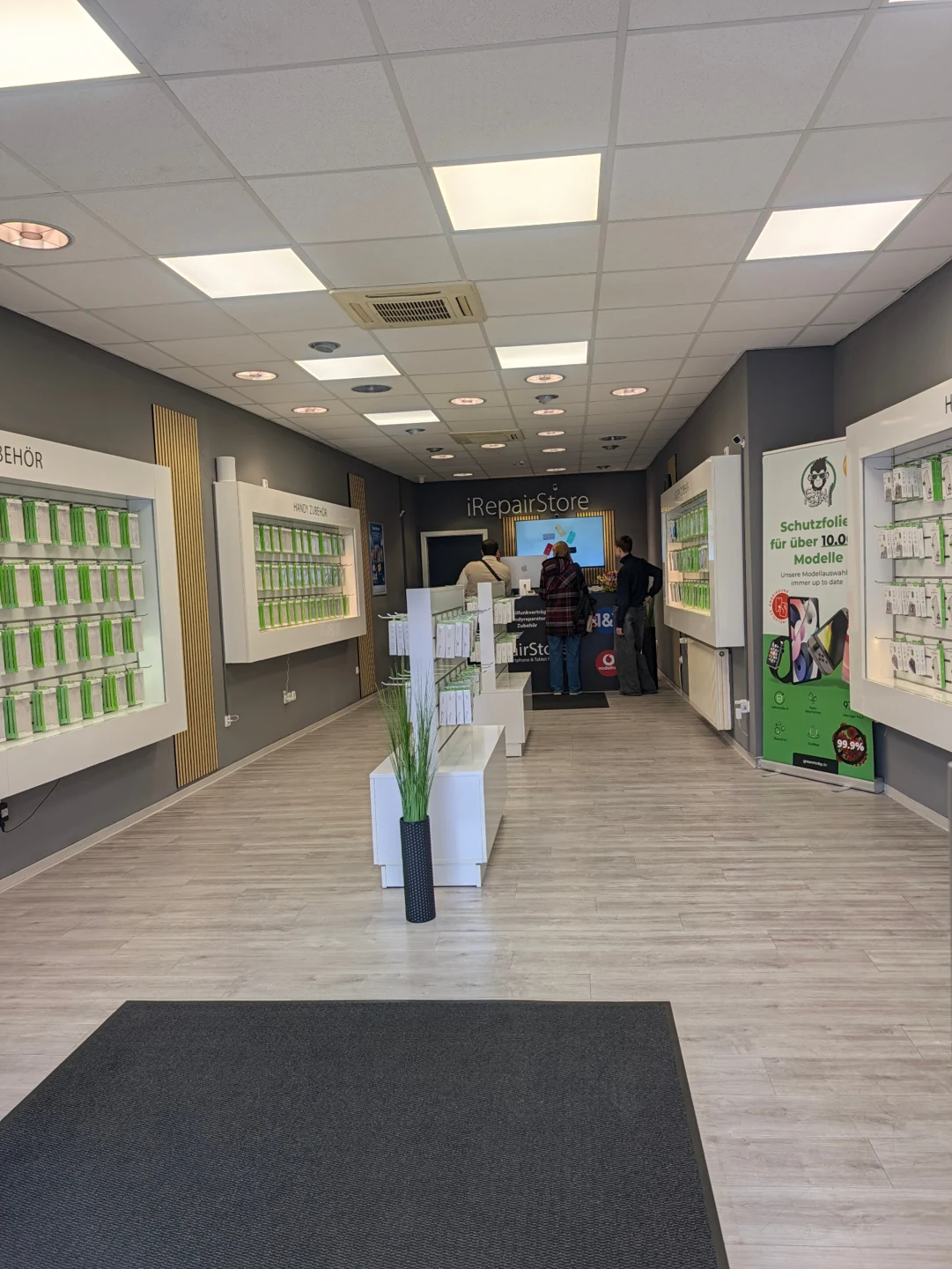 Innenansicht des iRepair Store in Bayreuth: heller Verkaufsraum mit Zubehörwänden voller Hüllen und Accessoires, Green Monkey Werbeaufsteller für Schutzfolien und Blick zur Beratungstheke mit iRepair-Store-Schriftzug.