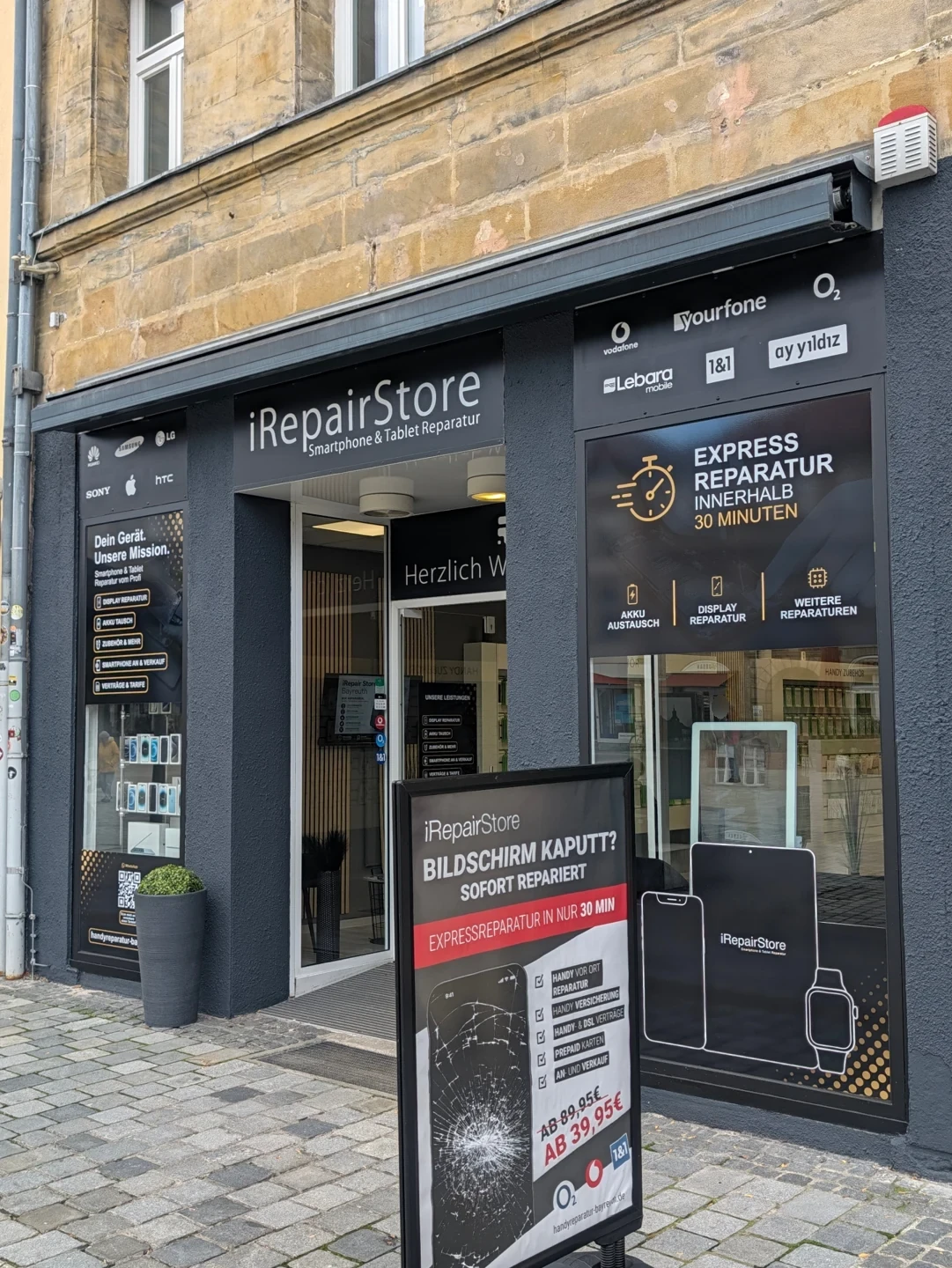 Schaufenster des iRepair Store in Bayreuth mit Aufsteller für Bildschirmreparaturen sowie Hinweisen zu Displayreparatur, Akkutausch, Handyverträgen und Mobilfunkanbietern.