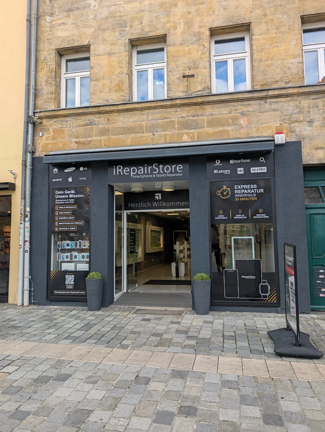 Eingang des iRepair Store in der Maximilianstraße 61 in Bayreuth mit Blick in den Laden, Handy-Zubehör an den Wänden und Smartphone-Motiven im Schaufenster.